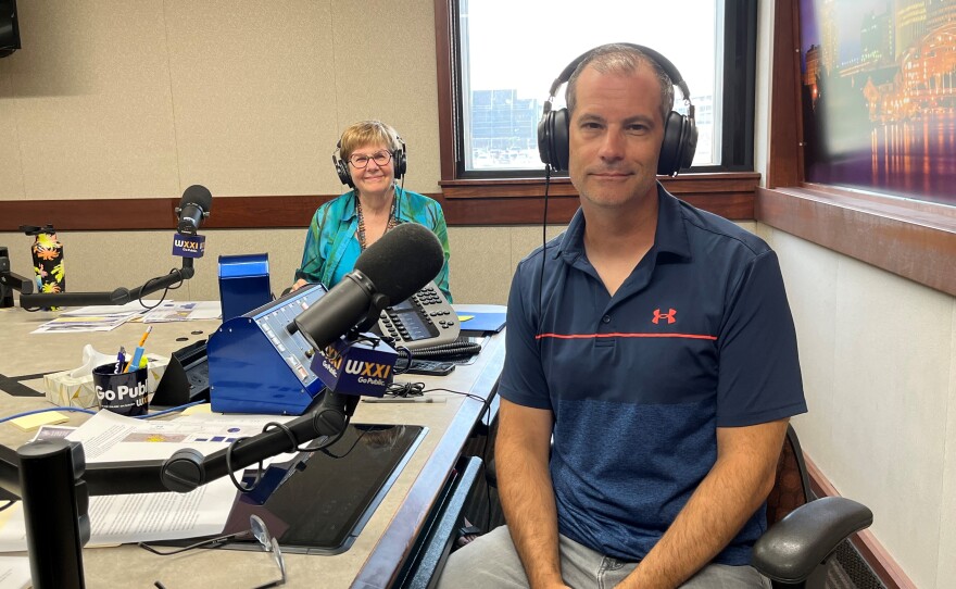 Two smiling people wearing headphones sit at a table in a radio talk studio: A woman at left has short blonde hair and is wearing a blue shirt and glasses; a man at right has short dark hair and is wearing a blue polo shirt.