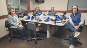 Four people wearing headphones sit at a table in a radio talk studio: a woman front left has very short grey hair and is wearing glasses, a blue and white blazer, white shirt, blue pants and blue shoes; a man front right has short dark hair and is wearing glasses, a navy puffy vest, white button-down shirt, jeans and sneakers; a woman back left has long brown hair and is wearing a black shirt; a bald man back right is wearing glasses, a grey turtleneck and a grey button-down shirt.