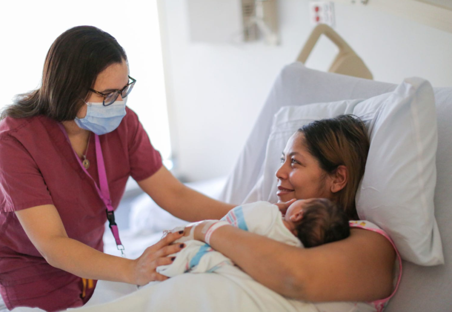A mother holds her newborn baby on the right, as her doula on the left, stands beside her hospital bed offering support.