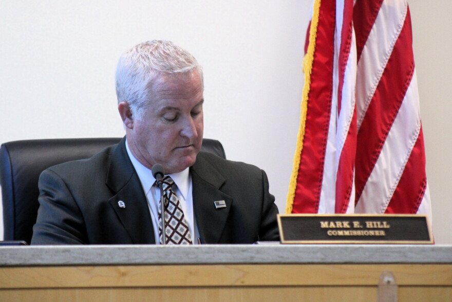 Commissioner Mark E. Hill sits at a desk with a nameplate in front of an American flag.