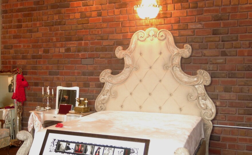 A white, ornate bed with upholstered head- and footboards with a small chandelier above and brick wall as a backdrop 