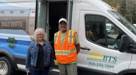A white woman and a black man smile side-by-side in front of a sprinter van. The van has a green alligator and the words "Shally Shuttle" painted on it.