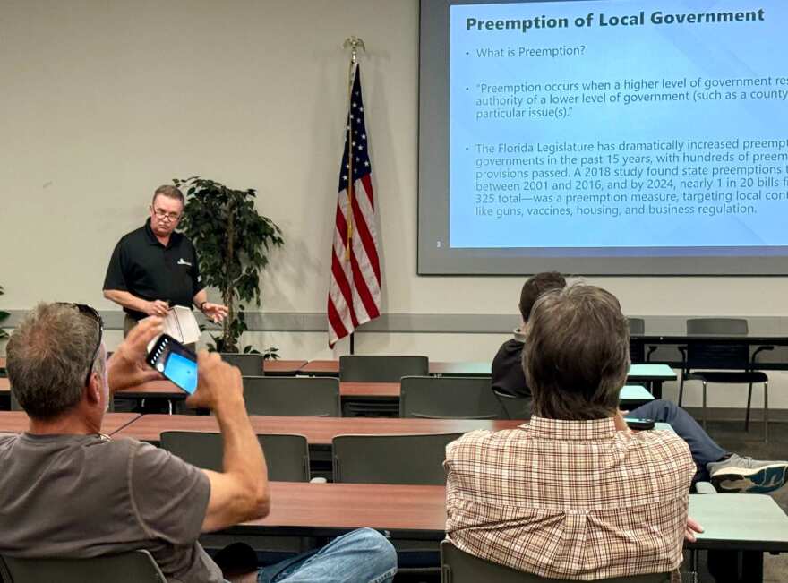 Rob Lewis in a room with three other people. On his right there is a flag of the United States of America and a a screen that says "Preemption of Local Government"