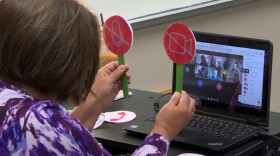 Tami McMahan sits in front of a computer on the first day of school, holding up icons representing the microphone and webcam buttons to help her virtual third grade class see whether they should turn theirs on or off.