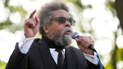 A man in sunglasses speaks into a mic held in one hand as he points at a high angle with the other. He's wearing a black suit jacket with a matching tie, along with a long sleeve white dress shirt with gold colored cufflinks. The background is heavily blurred, highlighting the speaker. 