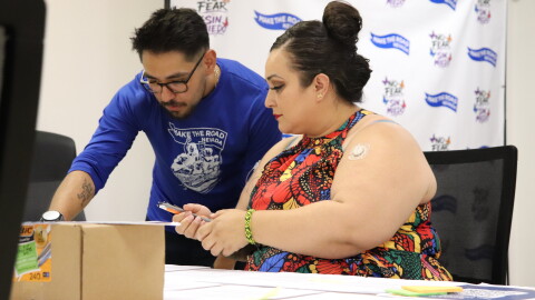 Erika Marquez sits a table looking down at something another person next to her is pointing out.