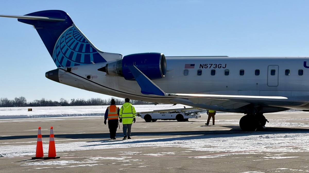 Two men dressed in neon yellow and orange walk towards the back of a United Airlines aircraft parked at the gate.