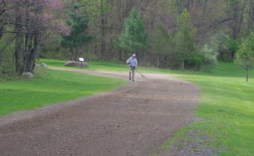 A man seen at a distance rakes a road with brownish gravel, in an area that is a mix of woods and lawn, in springtime