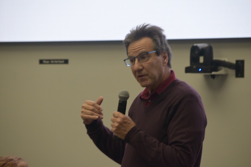 A man with a microphone, with glasses and wearing a maroon sweater.