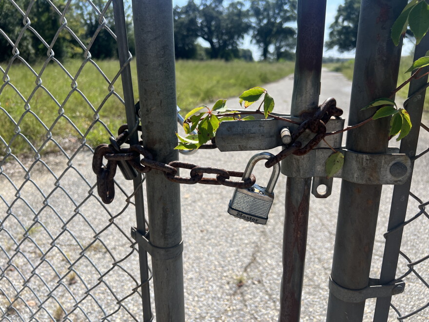 A padlock secures the gate at the Escambia Wood Treating Company Superfund site, where vines have grown around the fence and gate. An empty, overgrown lot is visible beyond the locked entrance.