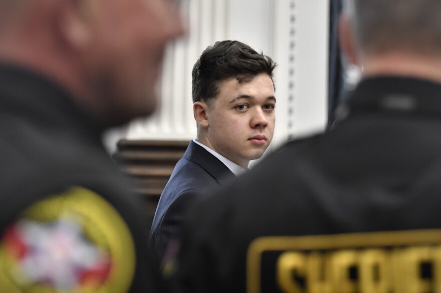 A teenager wearing a suit turns his head to look toward the camera, framed by the backs of two blurry security officers.