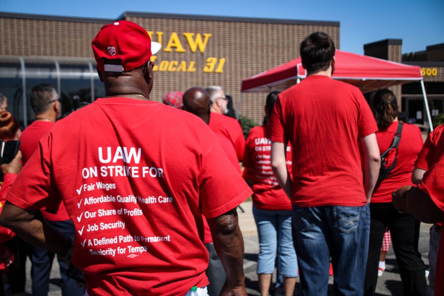 A man stands facing forward. The back of his shirt reads "UAW on strike for: fair wages, affordable quality health care, our share of profits, job security, a defined path to permanent seniority for temps."