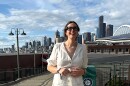 A smiling woman on a sunny day standing on a peer with the Seattle city scape in the background. She is wearing a white dress and dark rimmed glasses. 