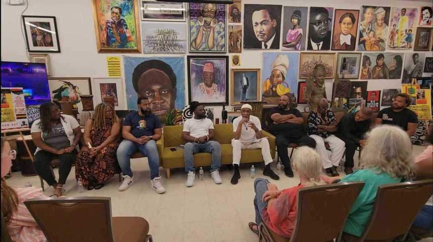 a group of African American women and men are seated on chairs and a sofa, behind artwork. In the center wearing white is community leader Ernest McMillan. 