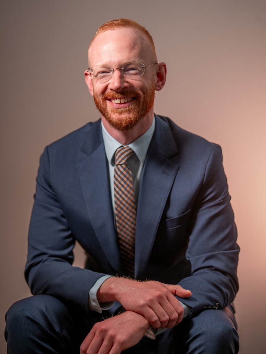 Man seated wearing a blue suit and patterned tie and smiling 