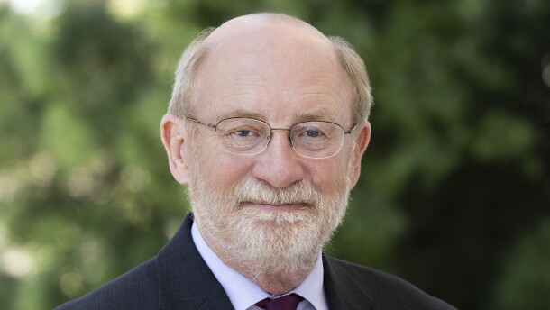 A bald man in a suit and dark purple tie looks into the camera. He is wearing glasses, a pin is on his right lapel. 