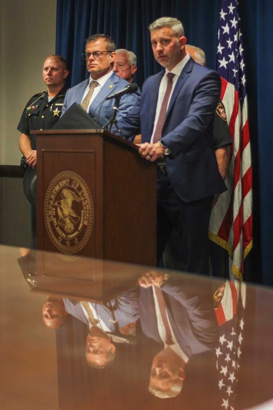 Two men in suits stand by a podium, with uniformed police behind them. They are reflected in a shiny table. 