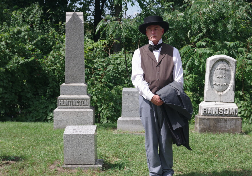 Man in vest with grey pants and bowtie, and top hat, and jacket over arm, poses next to stone markers
