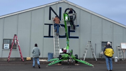 People paint "NO ICE" in black on the side of a building