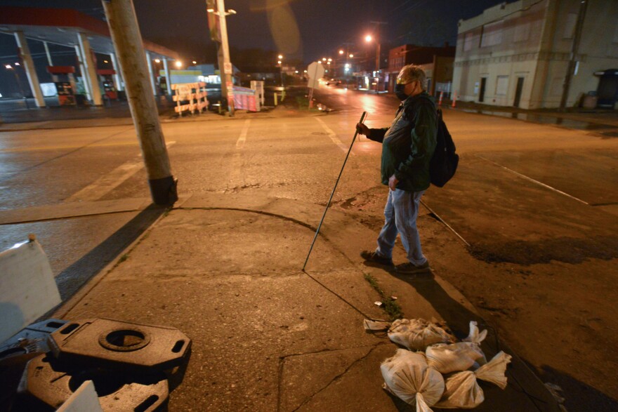 A man approaches a street corner while using a walking stick that indicates he is visually impaired. It is dark outside in the early morning.