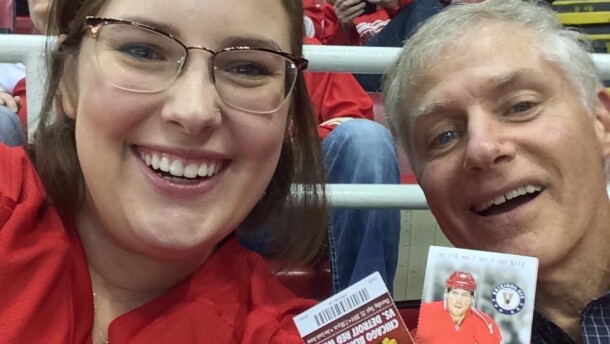 A selfie shows a young woman and an older man smiling in a stadium setting. Both individuals are looking directly at the camera, wearing Red Wings apparel. The woman wears glasses and has brown hair, while the man has short grey hair. The man holds up two printed hockey game tickets, showcasing logos and an image of a player. The ticket text reads "Chicago Blackhawks vs. Detroit Red Wings." The background features stadium seating with other spectators visible.