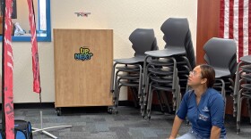 A young woman, sitting on the floor next to an open laptop and drone controller, watches a small drone float beneath a pink arch.