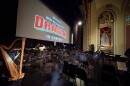 A photo taken from the wings of the Missouri Theatre stage, showing the empty chair setup for 80 musicians. A harp is featured prominently. A large projector screen with the opening of Dreamworks' 'How to Train Your Dragon' is above the percussion section.