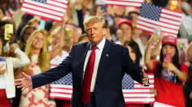 President Donald Trump stands with his arms outstretched. A crowd behind him waves American flags.