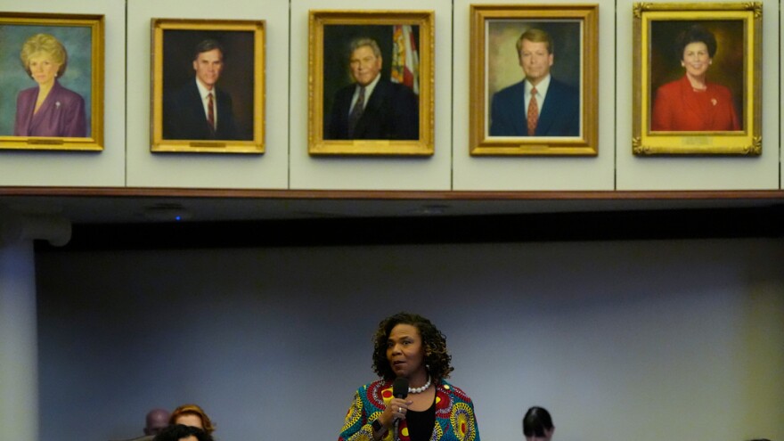 Black woman at the bottom of the photo speaking into a microphone with five portraits in gold frames on a wall above her