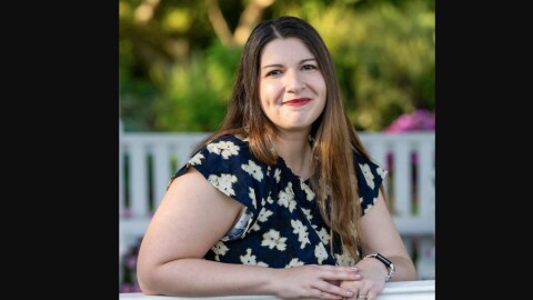 A woman in a blue dress with a flower pattern leans on a railing as she poses for a portrait photo with trees in the background. 
