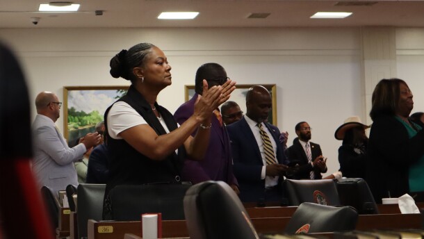 Rep. Carla Cunningham wears a black vest over a white short-sleeve shirt. Her hair is braided and in a bun. She is clapping, surrounded by other clapping lawmakers on the House floor.