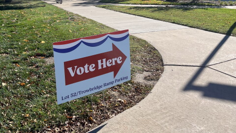 A red, white and blue Vote Here sign beside a campus sidewalk, with trees and a cyclist in the background.