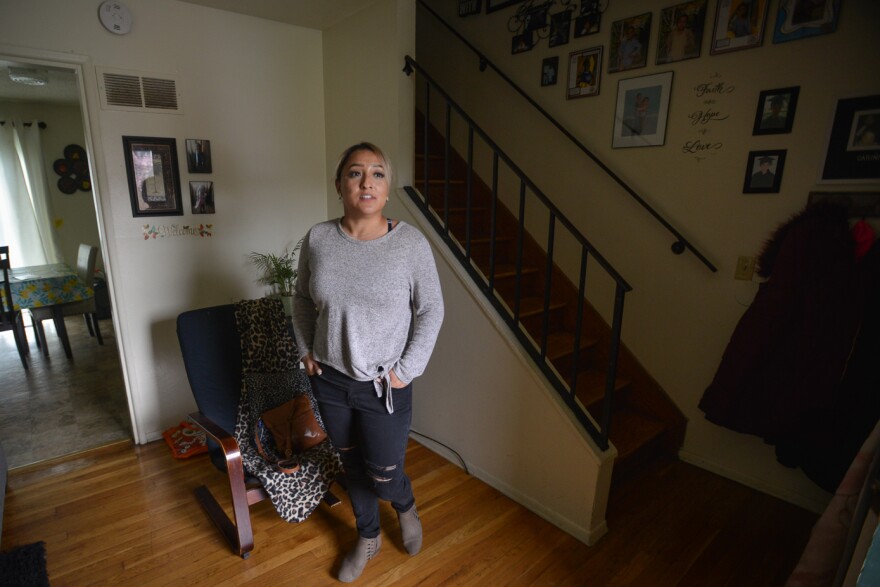 Libet Ojeda stands on a wood floor next to a set of stairs in a tidy home. On the wall behind her are many photos of her two children