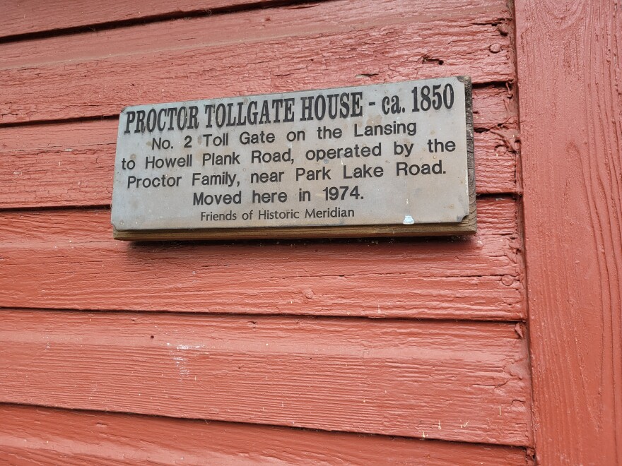 small metal sign on the red wood siding of a building. It reads, "Proctor Tollgate House- ca.1850. No. 2 Toll Gate on the Lansing to Howell Plank Road, operated by the Proctor Family, near Park Lake Road. Moved here in 1974. Friends of Historic Meridian"