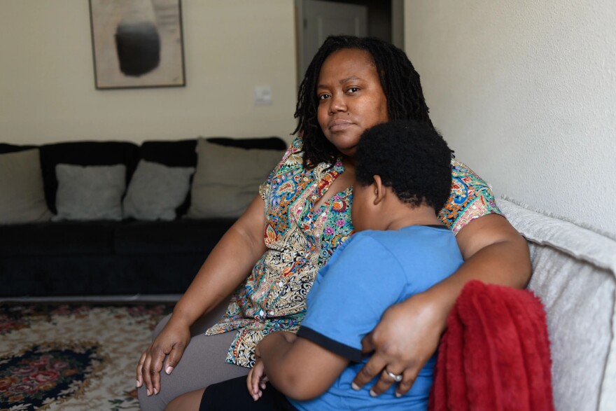 Black woman (mom) in print blouse holds a young Black boy in a light blue T-shirt, both sitting on a gray couch in a living room 