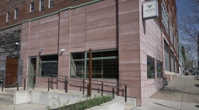Outdoor photo showing the exterior corner of a stone building. It has large, plate glass display windows. Some show kitchen items like a mixer and gourds in the display. Others allow a view into the restaurant. A small sign above reads "Anjin."