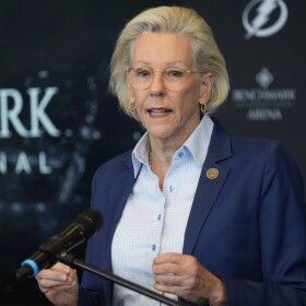 Woman with short blonde hair, glasses, blue blazer and blue shirt speaking into a microphone
