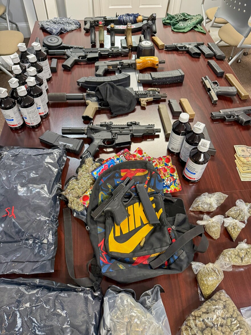  Multiple firearms, magazines, bottles, bags of plant material and cash are arranged across a table.
