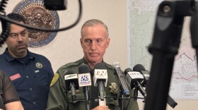 A sheriff standing up with several microphones pointed toward him. He is looking away from the camera.