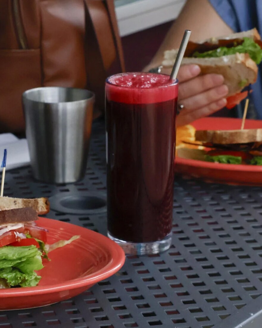 A glass of juice rests between two plates of sandwiches on a table.