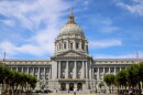 A view of the front of city hall with a few trees in the foreground and a blue sky with wispy clouds behind it. The city hall building is long and rectangular with columns in front, gold trim, and a big dome coming out of the center of it. You can see a few people walking in front of the building. 