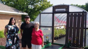 Two women stand with a man in front of a greenhouse filled with potted plants. From left to right, one woman wears a black dress with white and blue flowers decorating it, the man wears all black from his hat to his shorts, the last woman wears a bright red blouse and white pants. 