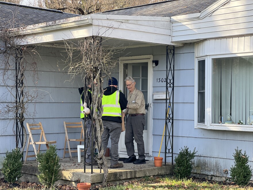 Two city workers in reflective vests stand on a porch and hand a brown paper bag to a man.