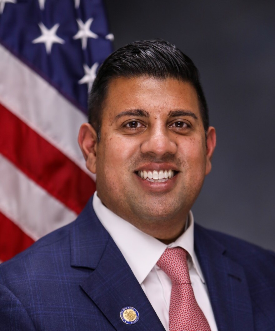 A man wearing a blue suit and red tie smiling in front of an American flag