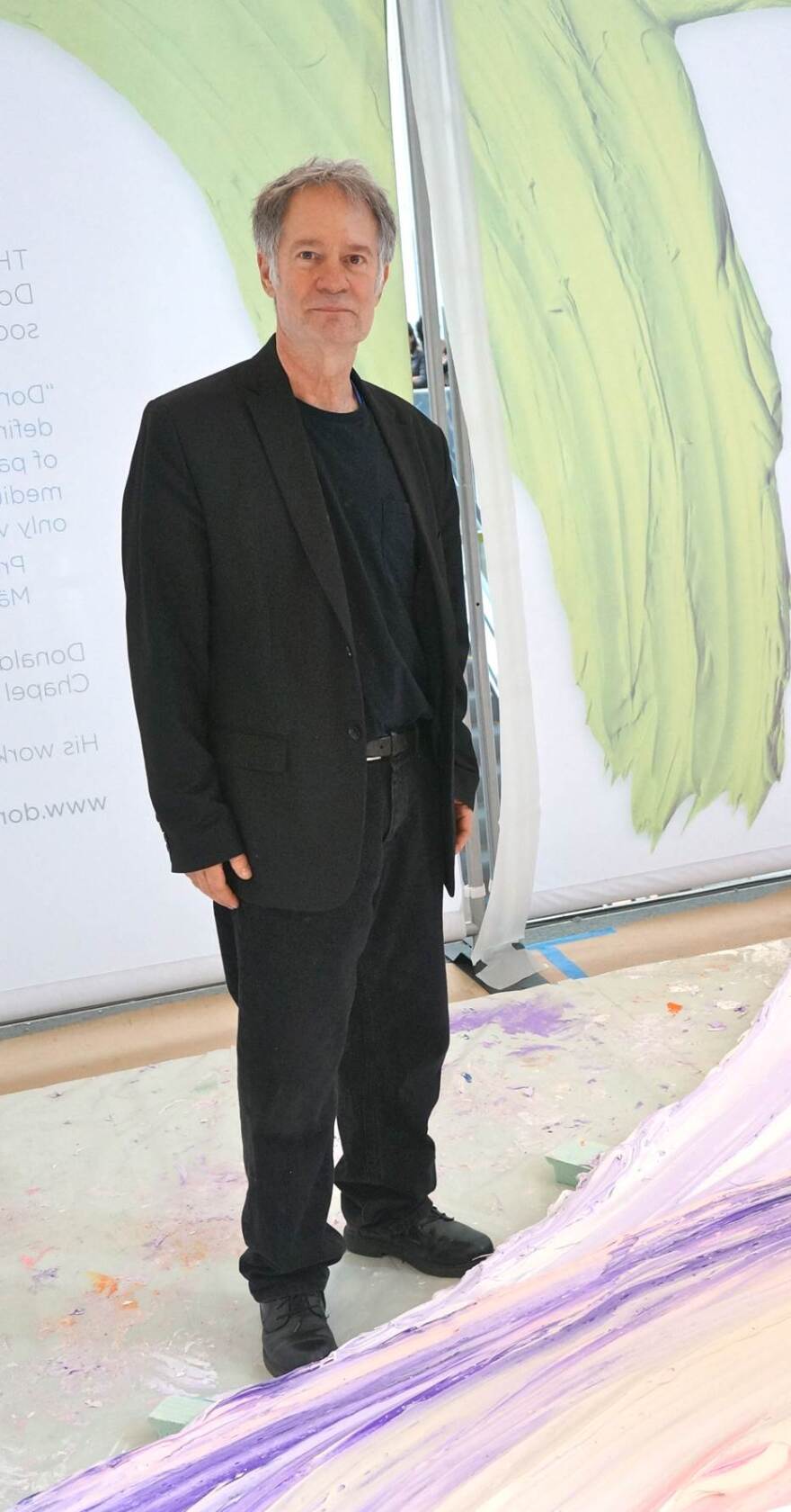 Martiny standing with the painting "Lenape." Behind him is the nylon curtain with a reproduction of one of his paintings on it.