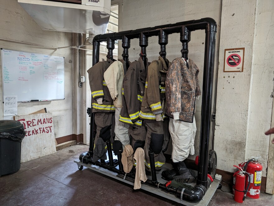 A Black PVC contraption with five hanging pipes where firefighting jackets and pants are hung. An air pump is hooked up to blow air into the pipes from the bottom.