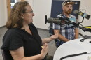 Two people sit in a radio studio talking at microphones. The person at left is gesturing while talking.