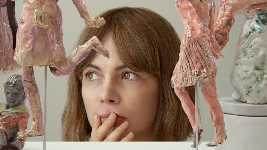 A woman looks at something out of frame as her head peeks above a tabletop with several small sculptures on it.