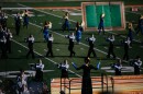 A drum major wearing a bow in her hair conducts marching performers on a football field. 