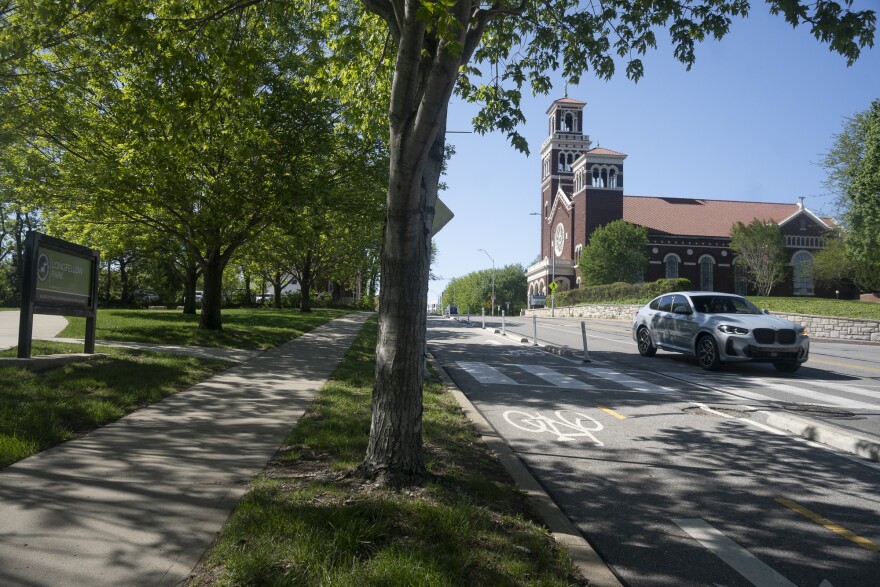 A shady park at left lies across a city street where a red, brick church can be seen in background at far right. A white car is traveling on the road near a bike lane.